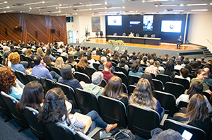 Imagem da Notícia Imagem de um auditório amplo, visto a partir do fundo da sala. Há centenas de pessoas sentadas em fileiras de cadeiras estofadas escuras, voltadas para a frente. O público é diverso, com homens e mulheres adultos, usando roupas sociais e casuais, como camisas, blusas e blazers. À frente, há uma mesa longa elevada, onde várias pessoas estão sentadas, compondo uma mesa de autoridades e palestrantes. Atrás delas, um grande painel com telas e logotipos é projetado na parede, indicando a realização de um evento institucional.