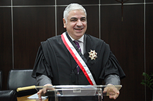 Imagem da Notícia O desembargador Samir Soubhia, homem de pele clara e cabelos curtos e grisalhos, está em pé atrás de um púlpito transparente. Ele usa uma toga preta e, sobre ela, uma faixa diagonal nas cores branca e vermelha, além de uma medalha ou insígnia presa ao lado esquerdo do peito. Sorri enquanto apoia as mãos no púlpito, onde há um microfone. Ao fundo, vê-se um ambiente interno formal, com parede escura e poltronas pretas.