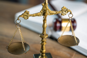 Imagem da Notícia Imagem em close de uma balança da justiça dourada, posicionada sobre uma mesa de madeira. A balança tem dois pratos metálicos suspensos por correntes finas, levemente inclinados, ligados a uma haste central ornamentada. Ao fundo, desfocado, há um livro grosso aberto ou apoiado, com aparência de livro jurídico, e um martelo de juiz (malhete) apoiado sobre ele. A iluminação é suave, destacando o brilho dourado da balança e criando um contraste com o fundo em tons neutros. A cena remete ao ambiente do direito, simbolizando justiça, equilíbrio e decisão.