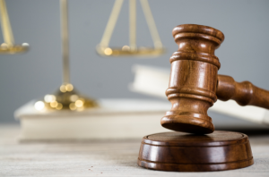 Ir para Imagem da Notícia A imagem mostra em primeiro plano martelo marrom em cima de mesa; e em segundo plano, desfocado, balança sobre livro