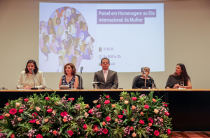 Imagem da Notícia Cinco pessoas estão sentadas à mesa de um evento no auditório do Fórum Ruy Barbosa, com um arranjo de flores coloridas à frente. No centro da mesa, o presidente do Tribunal Regional do Trabalho da 2ª Região (TRT-2), desembargador Valdir Florindo, veste terno cinza e gravata, em posição de destaque. Ao fundo, é projetado um slide com a arte do evento e os dizeres “Painel em Homenagem ao Dia Internacional da Mulher”. À esquerda e à direita do desembargador, quatro mulheres participam da mesa, compondo o painel. O ambiente é iluminado e formal, com foco na celebração do Dia Internacional da Mulher.