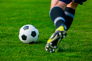 Imagem da Notícia Parte inferior das pernas de um jogador de futebol correndo atrás de uma bola em campo gramado