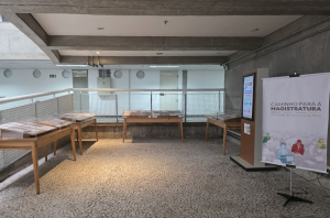 Imagem da Notícia Fotografia colorida, em plano geral, de um espaço de exposição montado em uma área de circulação interna, semelhante a um mezanino. O ambiente possui teto de concreto com vigas aparentes e chão revestido por mosaico de pedras portuguesas em tons claros.  À esquerda, dispostas diagonalmente, estão duas mesas de madeira clara com tampos inclinados, protegidas por caixas de acrílico transparentes, onde estão expostos documentos. Ao fundo, centralizada e encostada em um guarda-corpo metálico branco com tela de proteção, há uma terceira mesa de exposição similar às primeiras. Atrás do guarda-corpo, vê-se uma parede branca com janelas circulares tipo escotilha, uma caixa de luz cinza e uma porta de serviço bege.  À direita da imagem, em primeiro plano, encontra-se um banner vertical branco apoiado em um tripé preto. No topo do banner, lê-se o texto em destaque: "CAMINHO PARA A MAGISTRATURA", com uma linha fina abaixo e ilustrações coloridas na base representando silhuetas de pessoas. Ao lado do banner, um pouco mais ao fundo, há um totem digital vertical com a tela acesa exibindo informações.