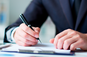 Imagem da Notícia Imagem de mãos masculinas usando terno escuro e assinando papel sobre a mesa