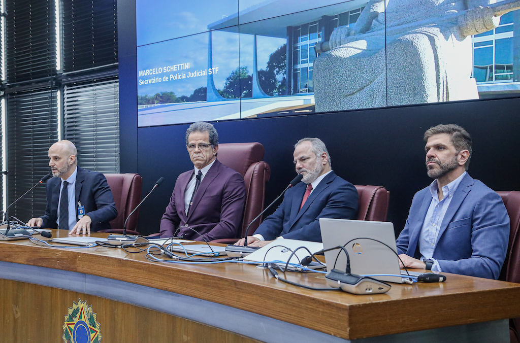 Imagem da Notícia Imagem em formato horizontal. Quatro homens estão sentados lado a lado atrás de uma mesa de madeira, em cadeiras altas. Todos usam ternos e estão voltados para a frente. Sobre a mesa há microfones, computadores, papéis e outros equipamentos. Na parte inferior esquerda, aparece parcialmente uma bandeira do Brasil. Ao fundo, há um grande painel com imagens projetadas e, à esquerda, uma tela vertical com informações.