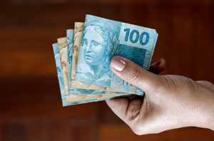 Imagem da Notícia Imagem de notas de 100 reais dobradas em mão feminina