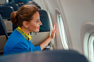 Imagem da Notícia Imagem em formato horizontal. Interior de uma aeronave. Uma mulher está sentada em uma poltrona próxima à janela. Ela usa uniforme azul com um lenço amarelo e verde no pescoço. Seu cabelo castanho está preso em um coque. A mulher está de perfil, voltada para a direita da imagem, com uma das mãos apoiada na janela oval do avião. As poltronas ao redor são azuis, com encostos altos. A luz externa entra pela janela, iluminando o rosto da mulher e parte do interior da cabine.