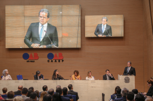 Imagem da Notícia No auditório da OAB/SP, autoridades estão sentadas à mesa no palco, diante de plateia. À direita, o presidente do Tribunal Regional do Trabalho da 2ª Região, desembargador Valdir Florindo, aparece em pé, no púlpito, discursando. Dois telões exibem sua imagem ampliada.