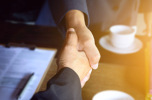 Imagem da Notícia Imagem em formato horizontal. Duas pessoas estão apertando as mãos. Apenas os braços e as mãos são visíveis. Ambas usam roupas formais em tons escuros. O aperto de mãos ocorre sobre uma mesa. Sobre a mesa há papéis com texto impresso, uma pasta ou prancheta e, ao fundo, uma xícara branca sobre um pires. A iluminação é quente, com uma faixa de luz amarelada no lado direito da imagem.