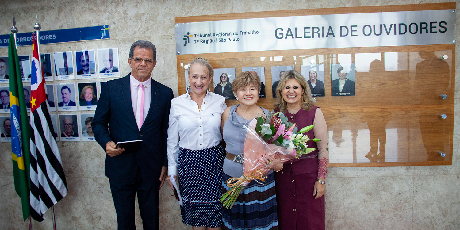 Quatro pessoas, incluindo uma mulher com buquê, posam sorridentes diante da Galeria de Ouvidores no Tribunal Regional do Trabalho em São Paulo.
