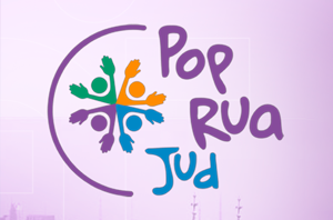 Ir para Imagem da Notícia Imagem em formato horizontal com fundo em tom claro, levemente rosado. À esquerda, há um elemento gráfico circular incompleto, formando quase um círculo. Dentro desse círculo, aparece um símbolo composto por quatro formas em cores diferentes (verde, laranja, roxo e azul), dispostas em cruz, com pequenos elementos que lembram folhas ou mãos estilizadas. À direita do símbolo, está o texto “Pop Rua Jud”, disposto em três linhas. As palavras estão em letras estilizadas e coloridas: “Pop” na parte superior, “Rua” no meio e “Jud” na parte inferior. As cores das letras variam entre roxo, azul e outras tonalidades. O fundo não apresenta outros elementos em destaque.