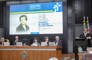 Ir para Imagem da Notícia Auditório do 20º andar do ed. Sede com mesa de autoridades à frente. Cinco pessoas estão sentadas atrás da bancada, enquanto o presidente do TRT-2, desembargador Valdir Florindo, fala em um púlpito à direita. Ao fundo, um telão exibe a foto da Desembargadora Neusenice de Azevedo Barretto Küstner, a homenageada que dá nome ao auditório. Bandeiras e elementos oficiais compõem o ambiente.
