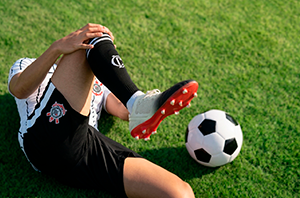 Imagem da Notícia Jogador de futebol caído em campo gramado, com mãos em um dos joelhos; há uma bola de futebol ao lado do homem