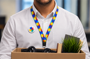 Imagem da Notícia Pessoa vestindo camisa social branca segura uma caixa de papelão com pertences de escritório, usando um cordão de pescoço e um broche no peito, ambos estampados com peças de quebra-cabeça coloridas (símbolo do espectro autista).