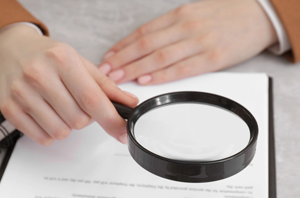 Imagem da Notícia Imagem de mãos femininas com lupa sobre documento em papel