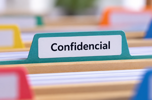 Imagem da Notícia Imagem em formato horizontal. Em primeiro plano, há uma divisória de plástico azul-esverdeada com uma etiqueta branca, na qual está escrito “Confidencial” em letras pretas. A divisória está encaixada entre pastas e documentos empilhados horizontalmente. Ao fundo, aparecem outras pastas e objetos coloridos, desfocados.