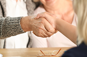 Imagem da Notícia Duas pessoas apertam as mãos sobre uma mesa; uma delas tem mão idosa, com rugas e veias aparentes