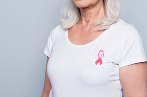 Imagem da Notícia Mulher de cabelos brancos na altura dos ombros, vestida com blusa branca e um laço rosa fixado na altura do peito esquerdo