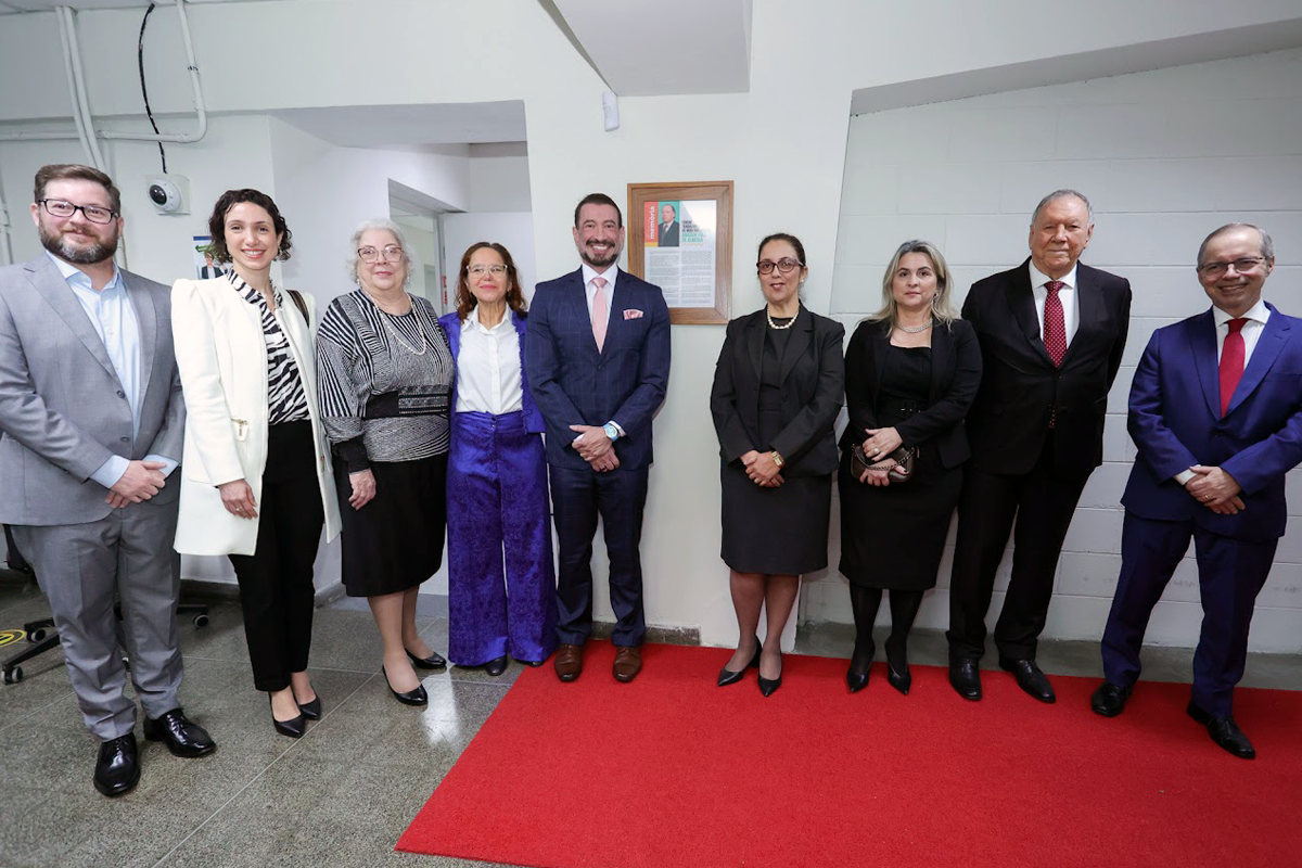 Grupo de pessoas em trajes formais posa lado a lado num evento interno, com um quadro pendurado na parede ao fundo e um tapete vermelho no chão