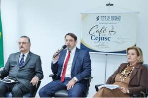 Imagem da Notícia Representantes do TRT-2 debatem estão sentados em cadeiras durante evento café com cejusc. Atrás, um banner exibe o nome do evento.