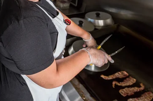 Imagem da Notícia Pessoa vista parcialmente e de costas, usando avental branco sobre roupa escura e luvas descartáveis. Ela segura uma pinça e manipula alimentos sobre uma grelha ou chapa. À direita, há pedaços de carne sendo grelhados. Ao fundo, aparecem utensílios e panelas sobre uma superfície de cozinha.