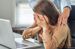 Imagem da Notícia Mulher sentada em frente a uma mesa com notebook, ela esconde o rosto com as duas mãos. Atrás da mulher, um homem em pé, com as duas mãos nos ombros dela.