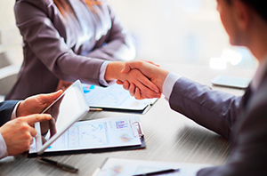 Imagem da Notícia Imagem em ambiente bem iluminado corporativo. Em primeiro plano, duas pessoas vestidas com roupas formais apertam as mãos sobre uma mesa, indicando um cumprimento profissional ou fechamento de acordo. Apenas os braços e as mãos são visíveis. À esquerda, outra pessoa, também com roupa social, segura uma prancheta com gráficos impressos e aponta para o documento com uma caneta. Sobre a mesa há papéis e um tablet. Ao fundo, levemente desfocadas, aparecem outras pessoas sentadas, sugerindo uma reunião.