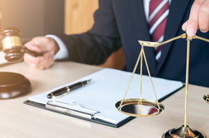 Imagem da Notícia Imagem em formato horizontal. Em primeiro plano, sobre uma mesa de madeira clara, há uma balança metálica com dois pratos suspensos por correntes. Ao lado da balança há uma prancheta com folhas de papel presas e uma caneta apoiada sobre ela. À esquerda da imagem, aparece um martelo de juiz (malhete) sobre a mesa. Ao fundo, parcialmente visível, está uma pessoa usando terno escuro e gravata listrada vermelha e branca, com uma das mãos próxima à balança.