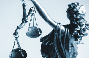 Imagem da Notícia Imagem em formato horizontal. Representação de uma escultura metálica da figura da Justiça. Aparece apenas a parte superior do corpo e o braço direito erguido. A figura segura uma balança com duas correntes, cada uma sustentando um prato circular. Os dois pratos estão suspensos e aparentam estar no mesmo nível. A escultura tem acabamento em tom cinza metálico. O fundo é claro, em tom branco ou azul muito claro, sem outros elementos visíveis.