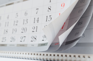 Imagem da Notícia A imagem mostra um calendário de parede, com várias páginas sendo folheadas. As folhas estão parcialmente levantadas, revelando os dias e números dos meses seguintes. Os números estão impressos em preto, e alguns dias aparecem destacados em vermelho.