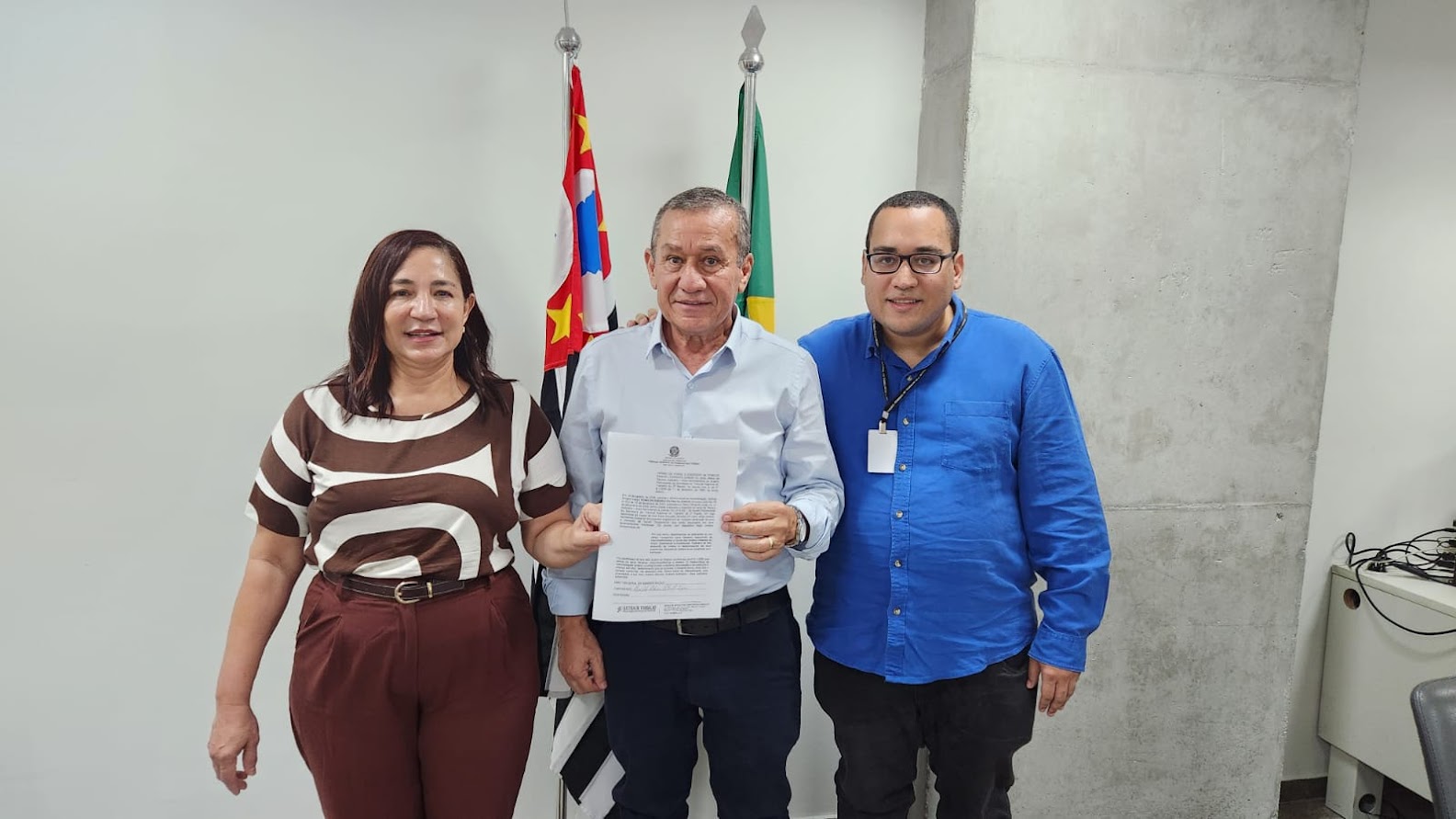 Uma fotografia em plano médio, capturada em um ambiente interno, mostrando três pessoas em pé, lado a lado, posando para a câmera. Ao fundo, veem-se duas bandeiras (do Estado de São Paulo e do Brasil) e uma parede de concreto aparente.