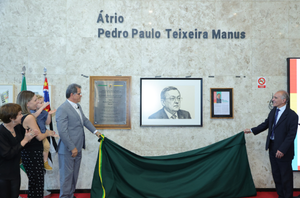 Imagem da Notícia Imagem em formato horizontal. Em uma parede clara, ao centro, há um quadro com um retrato em preto e branco de um homem, acima do qual está escrito “Átrio Pedro Paulo Teixeira Manus”. Ao lado do retrato há outros quadros e placas. À esquerda, quatro pessoas estão de pé diante da parede; entre elas, está o desembargador-presidente do TRT-2, Valdir Florindo, usando terno cinza. À direita, o desembargador Armando Augusto Pinheiro Pires, também de terno, segura uma extremidade de um tecido verde-escuro que cobre parcialmente a parte inferior da parede. Ao fundo, há mastros com bandeiras.