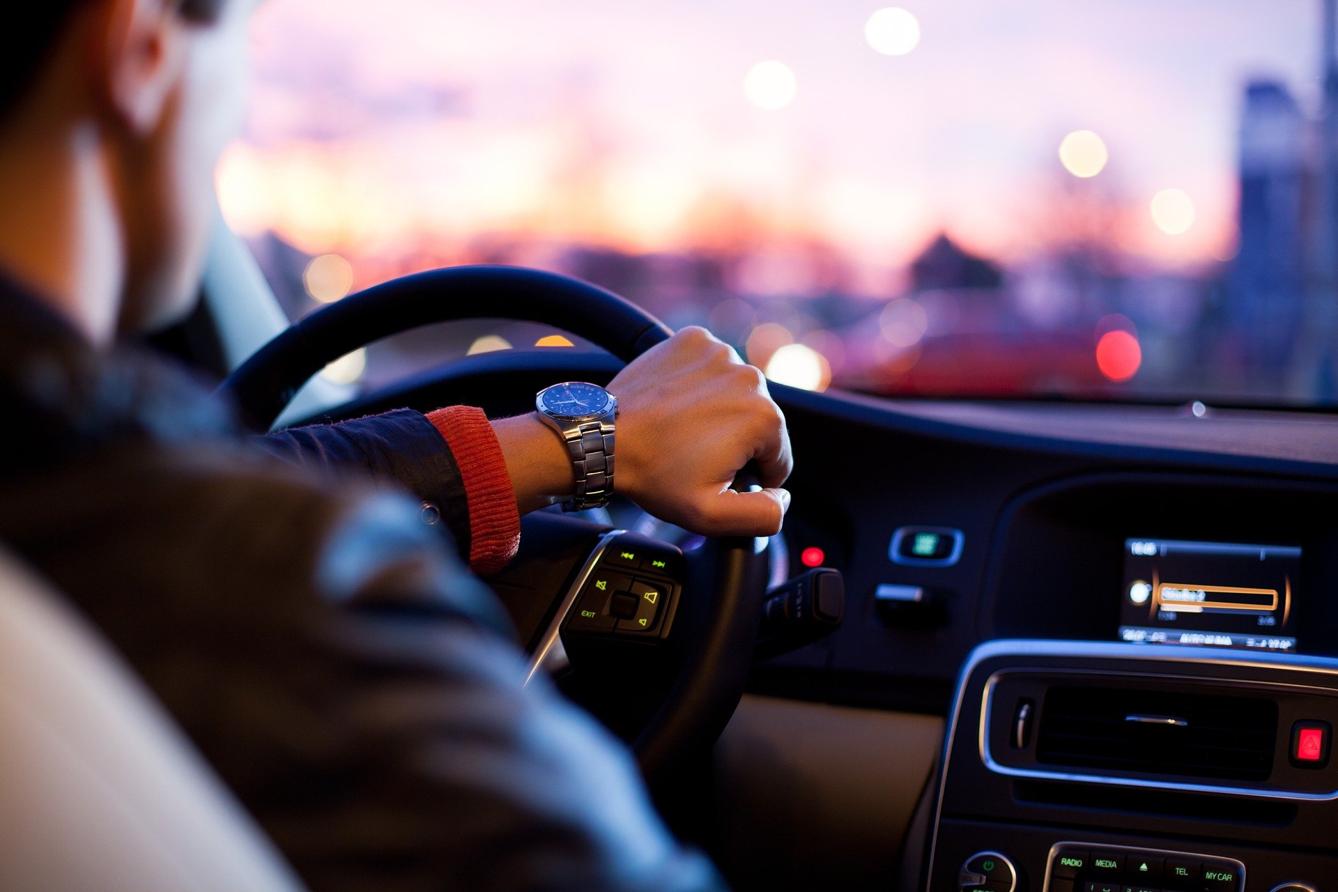 Homem branco de roupa preta segurando o volante
                    de um carro com mão esquerda. No pulso um relógio, e
                    à direita painel do carro com telas e botões.